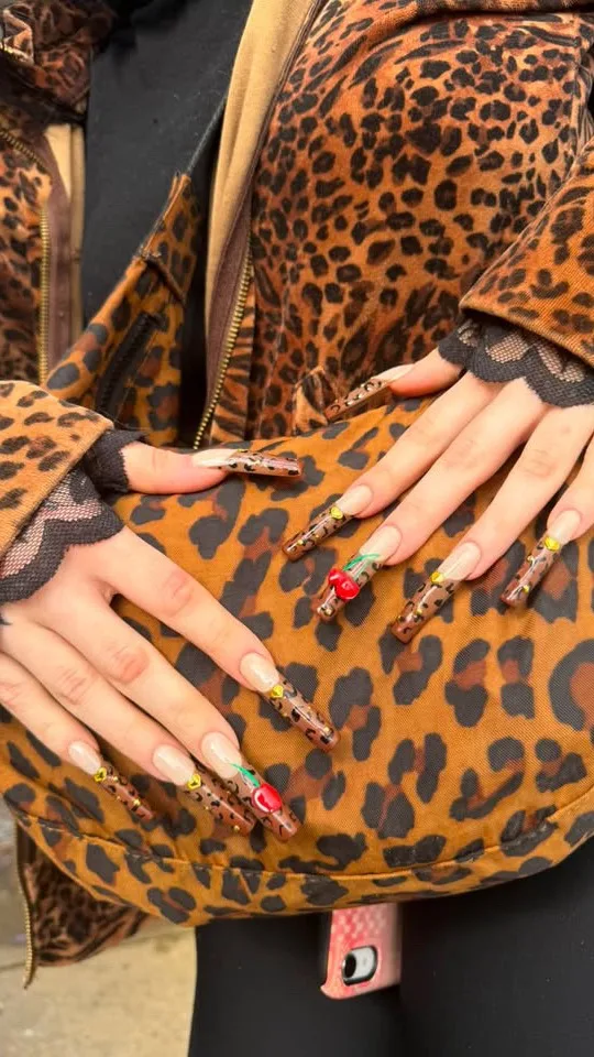 Long leopard-print nails with 3D cherries and gold gems, hands resting on a matching leopard jacket
