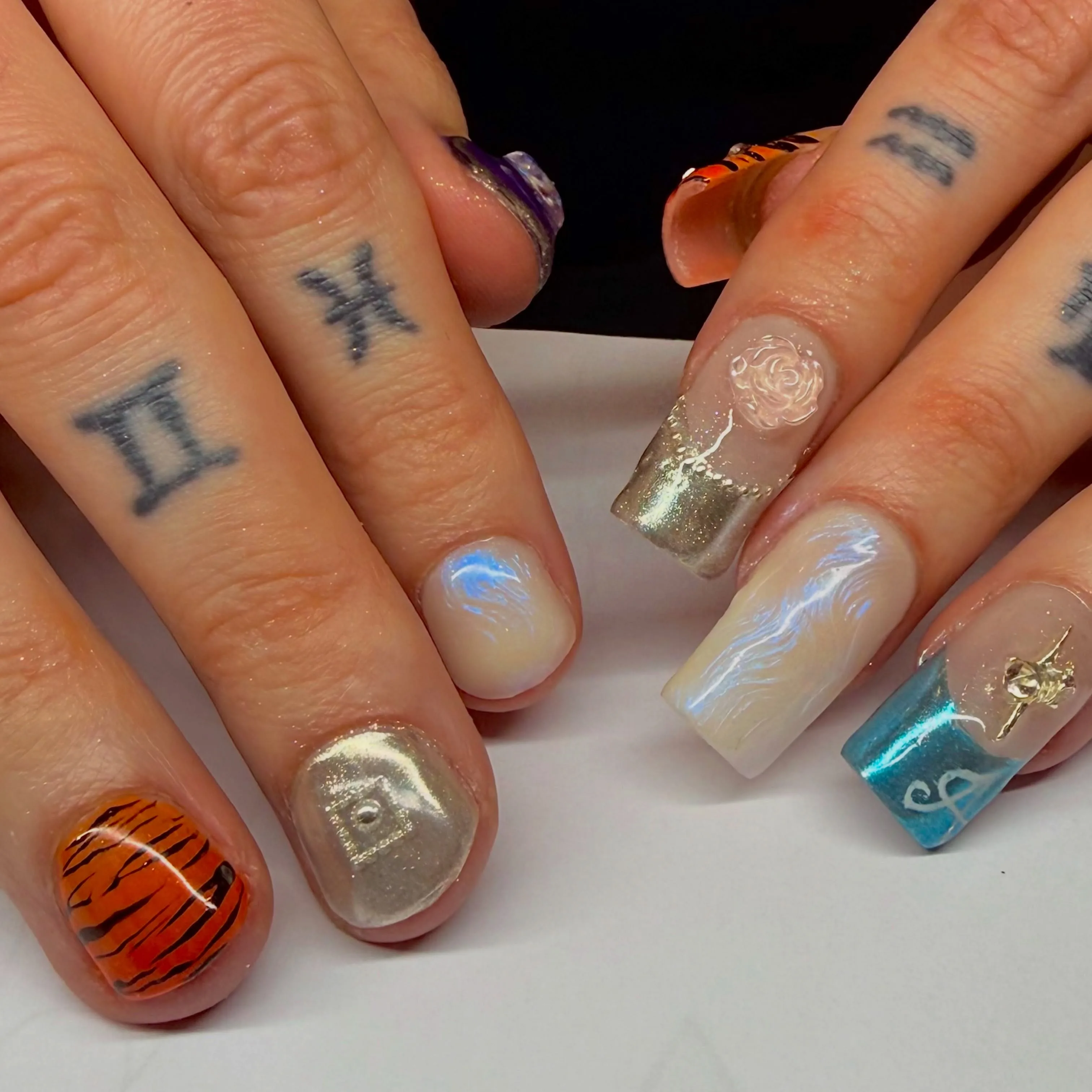 Tiger-stripe, gold chrome, opal and gem-set nail art on two tattooed hands with zodiac finger tattoos