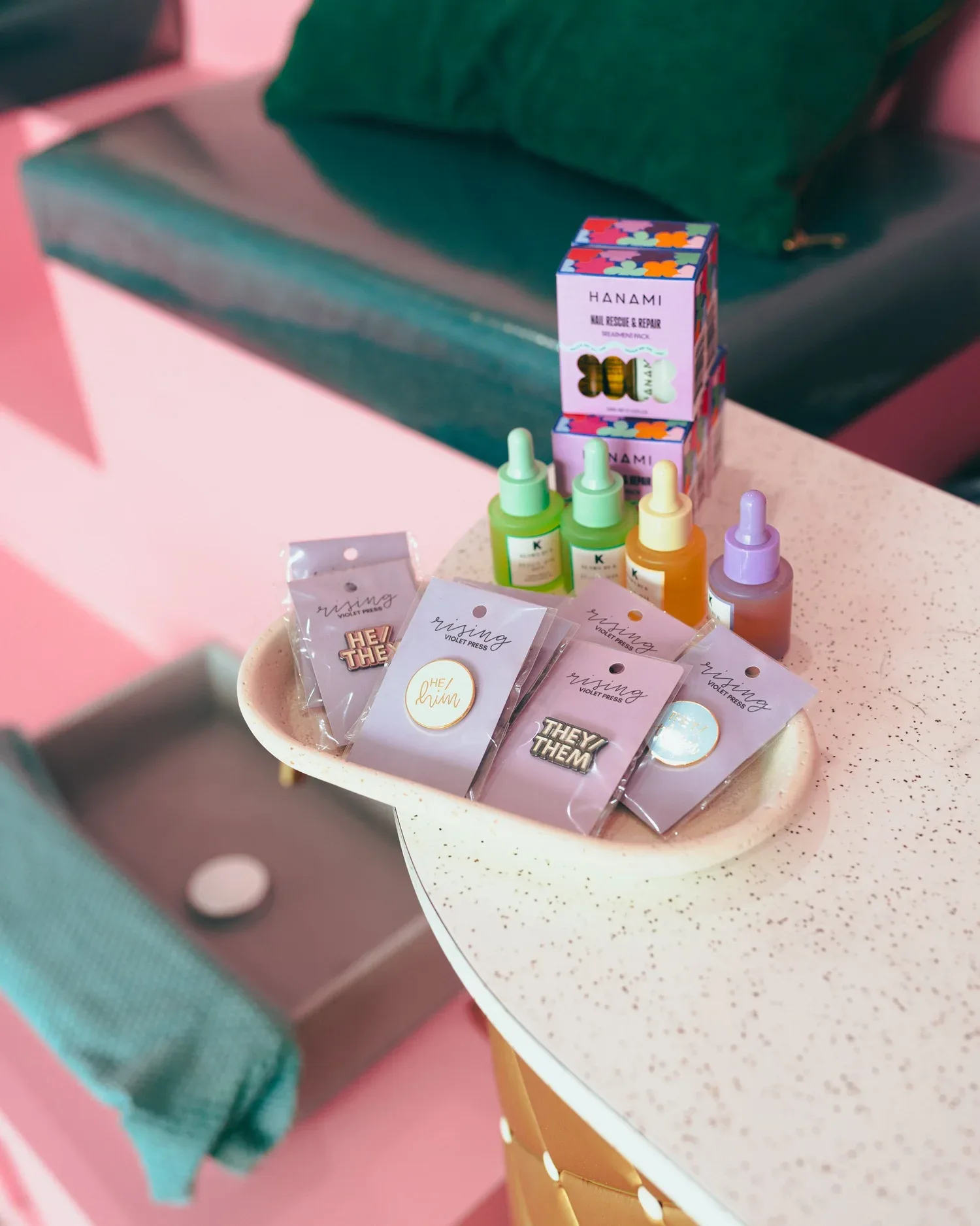 Retail display: pronoun pins (HE/HIM, HE/THEY, THEY/THEM) from Rising Violet Press in a ceramic dish, HANAMI Nail Rescue packs and pastel cuticle-oil droppers on a terrazzo table