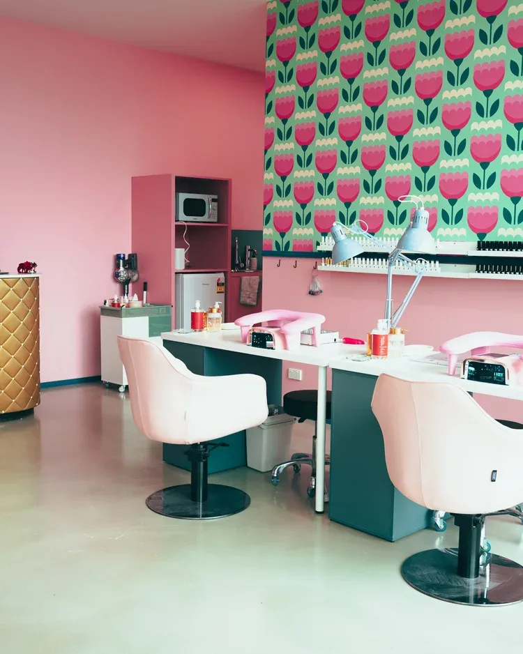The pink-and-mint salon interior — retro pink-tulip-on-mint feature wallpaper, two pink manicure chairs at a white bench, blue task lamps, and the gold quilted reception counter