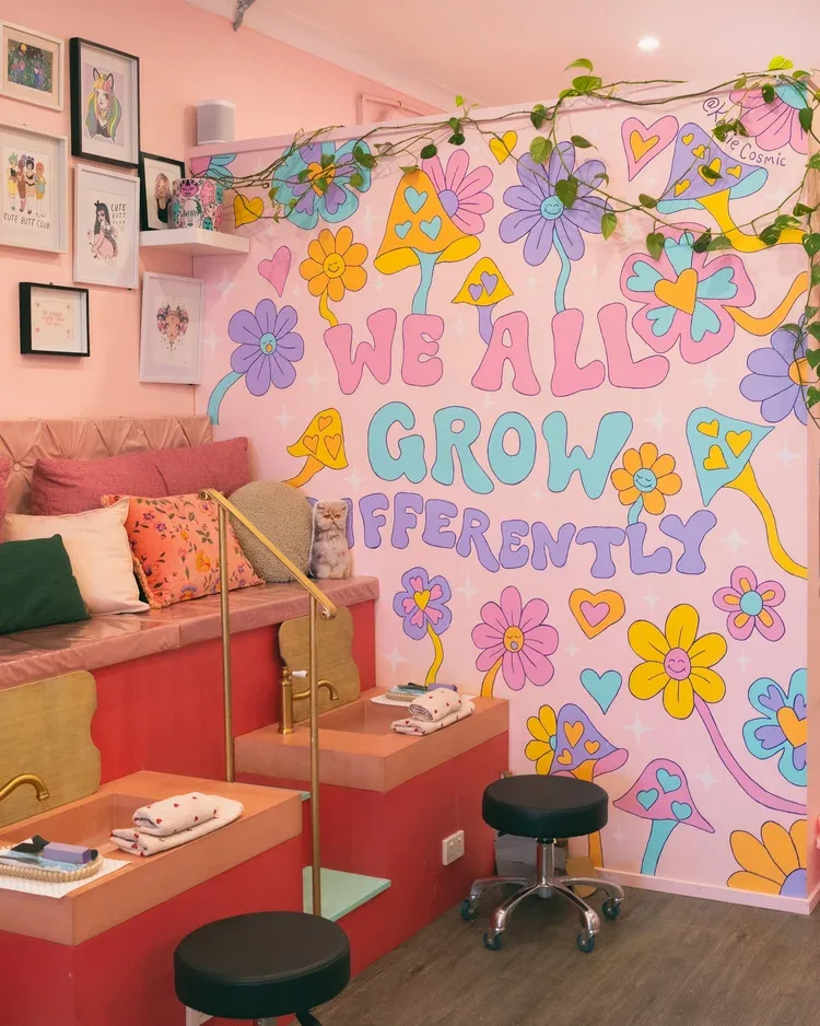 The pedicure bay — hand-painted 'WE ALL GROW DIFFERENTLY' groovy flower mural signed @cutie Cosmic, coral foot baths, and a gallery wall of queer art prints