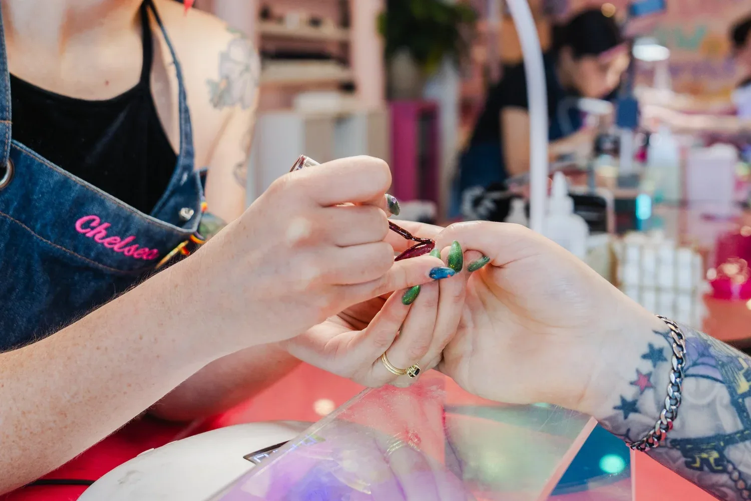 Close-up of a technician's hands painting a client's green and blue chrome nails, salon bokeh in the background
