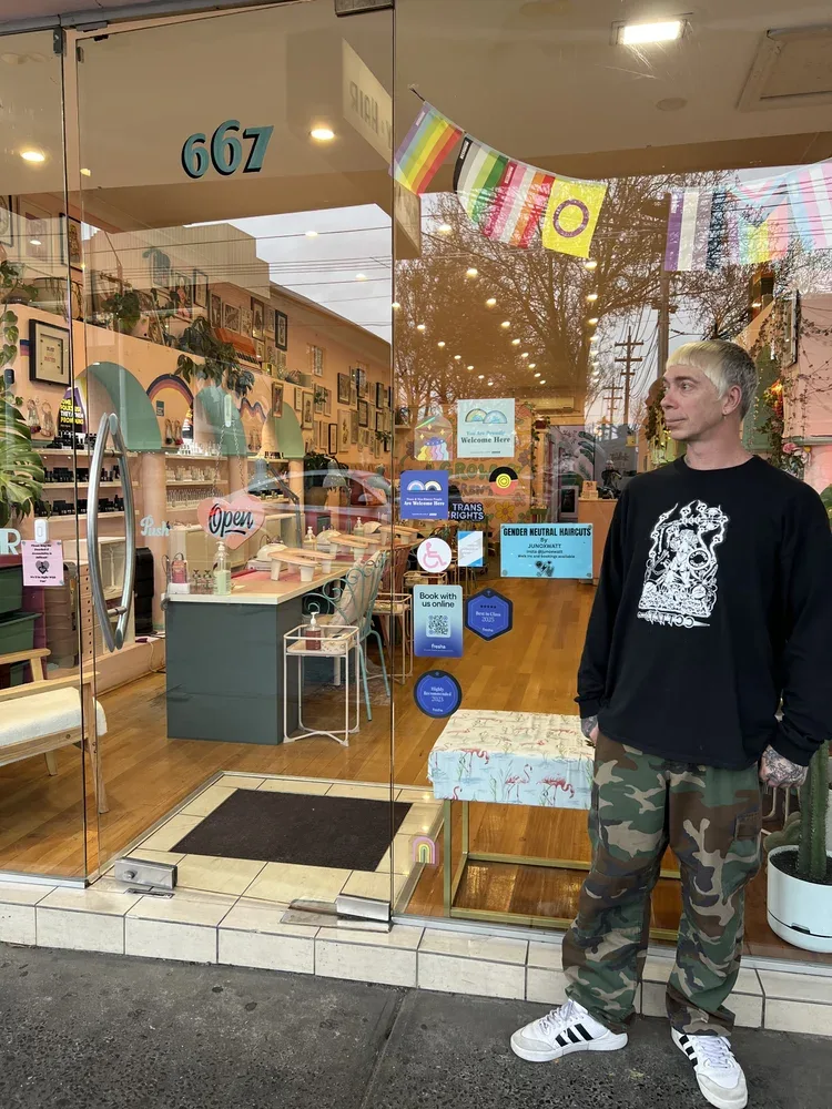 Juno standing outside the 667 High Street shopfront under rainbow bunting, beside signs reading 'GENDER NEUTRAL HAIRCUTS by JUNOXWATT', 'Trans Rights', and 'You Are Proudly Welcome Here'